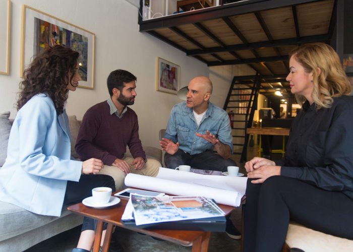 Interior designer presenting renovation project and discussing blueprint with clients. Group of people sitting on couch at home and talking. Renovation project concept